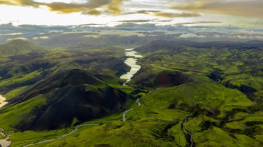 İzlanda 'nın yemyeşil vadileri, karanlık volkanik tepeler, dolambaçlı bir nehir ve uzak dağlar ve bulutlar tarafından çerçevelenmiş sakin bir göl..