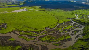 Örülmüş nehirler İzlanda ovalarında yeşil yosunlar boyunca akar. Volkanik topraklar ve arka planda görünen uzak dağlar vardır..