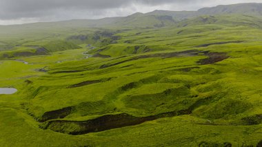 İzlanda 'da uçsuz bucaksız yeşil bir manzara. Yuvarlak tepeler, dolambaçlı vadiler ve dereler. Bulutlu gökyüzü volkanik araziyi aydınlatıyor..