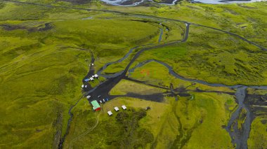 İzlanda 'da, yosunlu volkanik ovalar, çakıl yolları ve uzak akarsularla çevrili yeşil çatılı çadırları olan bir kamp alanının hava görüntüsü..