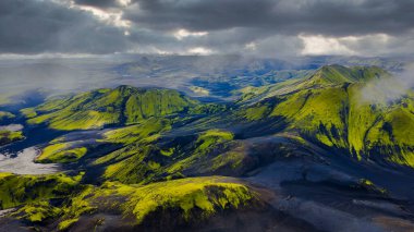 Yeşil yosun, engebeli dağlar, dolambaçlı bir nehir ve karamsar bir atmosfer yaratan bulutlu İzlanda volkanik arazisinin havadan bakış açısı.