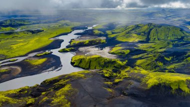 Sarmal bir nehir, yeşil yosun kaplı tepeler, siyah kum ve bulutlu gökyüzü altında engebeli oluşumlar içeren İzlanda 'nın volkanik arazisinin havadan perspektifi.