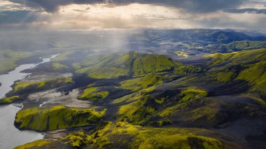 Yeşil yosun kaplı tepeler, karanlık toprak, dolambaçlı bir nehir ve bulutları aşan güneş ışığından oluşan İzlanda 'nın volkanik arazisinin hava perspektifi..