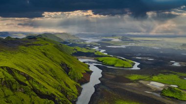 İzlanda 'nın volkanik arazisinin hava perspektifi kıvrımlı bir nehir, kara toprak, yeşil yosun kaplı tepeler ve bulutları aşan güneş ışığı..