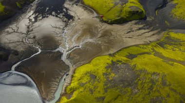 Örgülü nehir, İzlanda 'da yeşil yosun kaplı araziyle karanlık volkanik topraklar boyunca akar. Kırmızı bir yapı engebeli araziye ölçek ekler..