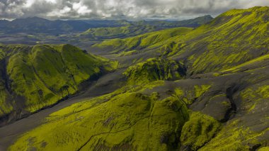İzlanda 'nın volkanik arazisinin hava perspektifi, siyah kayalar üzerinde yeşil yosunlar, kayan tepeler, derin vadiler ve bulutlu bir ufuk..