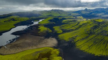 İzlanda 'nın volkanik arazisinin hava perspektifi yosun kaplı tepeler, dolambaçlı bir nehir, engebeli dağlar ve arka planda bulutlu bir gökyüzü içerir..