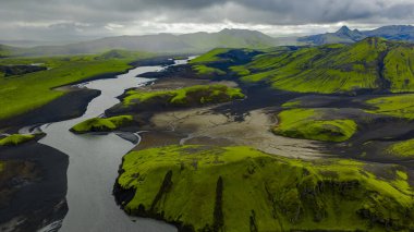 Sarmal bir nehir yosun kaplı tepeler ve karanlık volkanik topraklar boyunca akar, arka planda sivri tepeler İzlanda 'da bulutlu bir gökyüzünün altında.