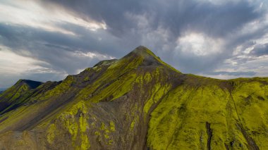 İzlanda 'da volkanik bir dağın dramatik hava manzarası, canlı yeşil yosunlar, karanlık kayalık arazi ve bulutlu gökyüzünde kırılan güneş ışığı..