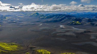 İzlanda 'nın volkanik dağlarının hava perspektifi; siyah kum düzlükleri, kıvrımlı nehirler, yeşil yosun yamaları ve bulutlu bir gökyüzünün altında engebeli dağlar..