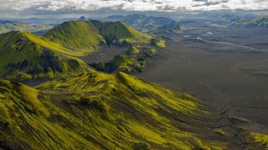 İzlanda 'nın volkanik arazisinin hava perspektifi, yeşil yosun kaplı dağlar, karanlık volkanik kum, dolambaçlı bir nehir ve bulutlu bir ufuk..