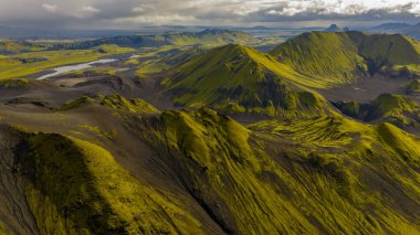 Yeşil yosun kaplı dağları, karanlık toprağı ve parçalı bulutlu gökyüzünün altındaki dolambaçlı bir nehri olan İzlanda 'nın volkanik dağlarının hava perspektifi..