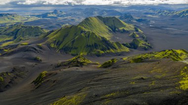 Yeşil yosun kaplı dağlar, karanlık toprak, yuvarlanan tepeler ve parçalı bulutlu bir gökyüzünün yer aldığı İzlanda 'nın volkanik arazisinin geniş hava manzarası..