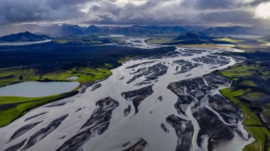 Örülmüş nehirler, İzlanda 'daki karanlık volkanik topraklar ve yeşil yosunlar boyunca akar. Bulutlu bir gökyüzünün altında küçük bir göl ve engebeli dağlar sahneyi tamamlıyor..