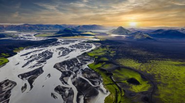 İzlanda 'nın örülmüş nehirlerinin hava perspektifi karanlık volkanik topraklar boyunca akar, yeşil yosunlar ve altın bir günbatımı altında uzak konik dağlar.