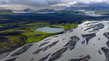 Örülmüş bir nehir, İzlanda 'daki karanlık volkanik arazide akmaktadır, buna karşın yeşil bir yama ve göl vardır. Bulutlu bir gökyüzünün altında yükselen engebeli dağlar.