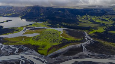 Karanlık toprak, yeşil yosun, örgülü nehirler, dingin bir göl ve alçak bulutların altındaki engebeli dağlar İzlanda 'nın volkanik arazisinin havadan bakış açısı..