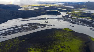 Sarmal nehirleri, yeşil yosunları, kara toprağı, dağınık tepeleri ve ufukta soluk bir gökkuşağı olan İzlanda 'nın volkanik manzarasına hava perspektifi..