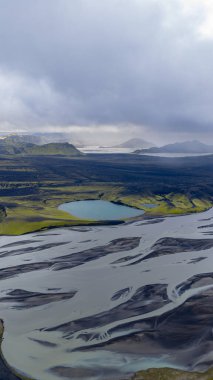 Örülmüş nehir, yeşil yosun kaplı tepeler, mavi bir göl ve İzlanda 'da bulutlu bir gökyüzünün altındaki uzak dağlarla karanlık volkanik arazide akar..