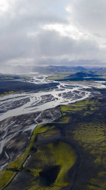 Örülmüş nehirler, İzlanda 'da bulutlu bir gökyüzünün altındaki uzak dağlarla çevrili yeşil yosunlarla kaplı kül arazisinden akar..