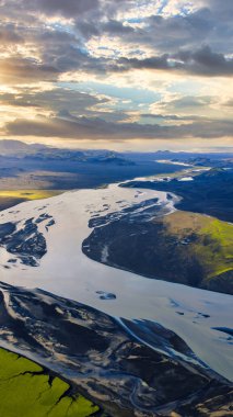 Dolambaçlı bir buzul nehri, altın güneş ışığı altında yeşil yosunlarla karanlık volkanik arazide akar. Kar kaplı dağlar uzaklardan görülebilir..