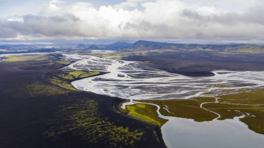 Örgülü nehir sistemi, İzlanda 'da bulutlu bir gökyüzünün altında yeşil yosun yamaları, gümüş su yolları ve uzak dağlarla volkanik arazide akar..