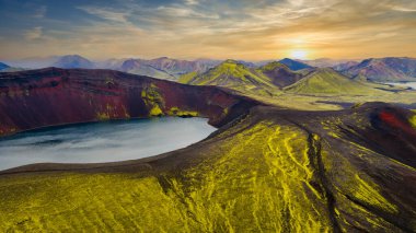Yeşil yosunlarla çevrili kırmızımsı kahverengi duvarları olan volkanik bir krater gölü. Arka planda engebeli volkanik dağlar ve altın bir günbatımı görünüyor..