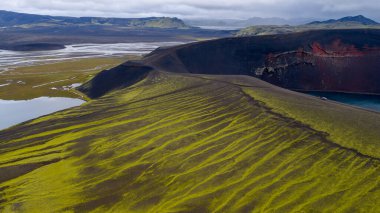 Yeşil yosun, krater gölü, dolambaçlı nehirler, geniş düzlükler ve bulutlu gökyüzünün altındaki uzak dağlar ile İzlanda 'nın volkanik arazisinin havadan görünüşü..