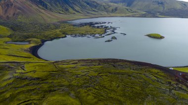 Yeşil yosun kaplı volkanik arazi, engebeli dağlar ve araçlı görünen toprak yollarla çevrili küçük bir adası olan göl manzarası..