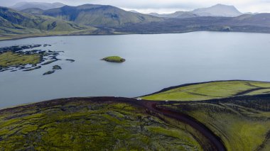 Etrafı yosun kaplı volkanik arazi, engebeli dağlar ve İzlanda 'nın dolambaçlı toprak yolu ile çevrili otlak bir adası olan sakin bir göl manzarası..