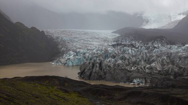 Mavi ve beyaz buzlu sivri buzullar karanlık volkanik kayalarla çevrili bulanık bir göle uzanır. Geniş ölçekte iki şekil vurgulanıyor..