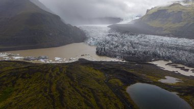 İzlanda 'da bir buzul yeşil yosunlu volkanik manzaralarla çevrili çökelti dolu bir lagüne doğru uzanır. Ön planda yansıtıcı bir gölet görülebilir..