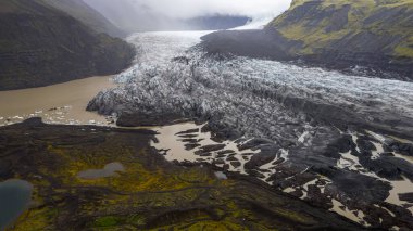İzlanda 'da bir buzul, koyu volkanik kayalar, yeşil yosun kaplı tepeler ve sisli uzak dağlarla çevrili çamurlu bir buzul gölüne akar..