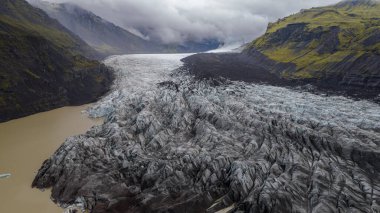 Bir buzulun sivri buz mavisi yarıkları volkanik arazi ve alçak bulutların altındaki yosunlu dağlarla çevrili çamurlu bir buzul gölüne yol açar..