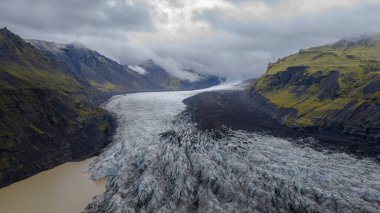 İzlanda 'da bir buzulun volkanik dağlarla kaplı yosunların arasında aktığı derin yarıklar, bir buzul gölü ve alçak bulutlar zirveleri kaplıyor..