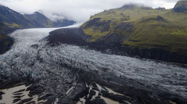 Derin yarıklara ve volkanik küllere sahip bir buzulun yosun kaplı dağlar arasında aktığını, alçak bulutların İzlanda 'nın engebeli manzarasında tepeleri gizlediğini..