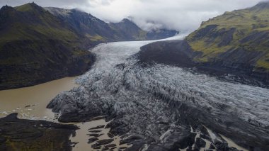 İzlanda 'daki bir buzulun karanlık volkanik dağlar arasında uzanan yarıklar ve kül çizgileri vardır. Kenarlarında buz parçaları olan çamurlu bir buzul nehri..