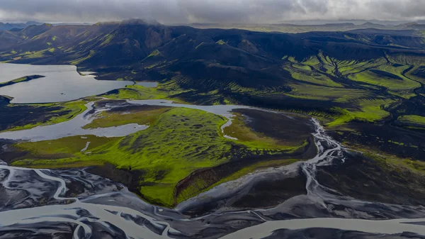 Karanlık toprak, yeşil yosun, örgülü nehirler, dingin bir göl ve alçak bulutların altındaki engebeli dağlar İzlanda 'nın volkanik arazisinin havadan bakış açısı..