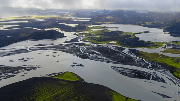 Örülmüş nehirlerin yeşil yosunlarla çevrili karanlık volkanik topraklar boyunca uzanan havadan bakış açısı. Arka planda göller ve sisli dağlar var..
