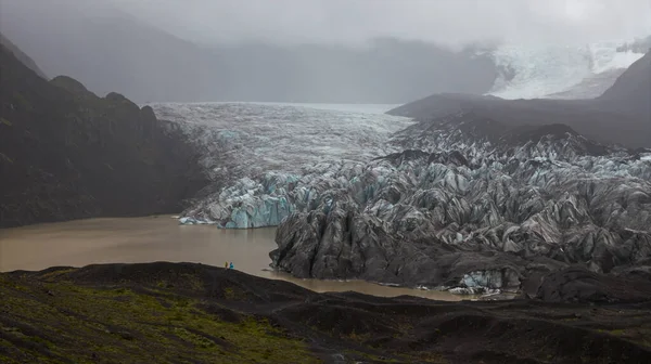 Mavi ve beyaz buzlu sivri buzullar karanlık volkanik kayalarla çevrili bulanık bir göle uzanır. Geniş ölçekte iki şekil vurgulanıyor..