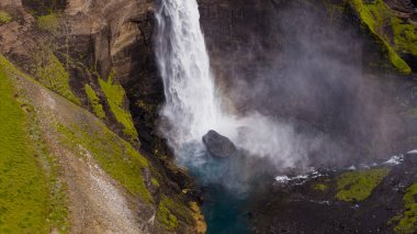 Güçlü şelale, volkanik kaya, yeşil yosun ve İzlanda 'nın engebeli arazisindeki üsten yükselen sisle çevrili turkuaz bir havuza dalıyor..