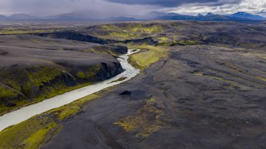 İzlanda 'nın volkanik arazisinin hava perspektifi kıvrımlı bir nehir, yeşil yosun yamaları, engebeli dağlar ve uzakta bulutlu bir gökyüzü..