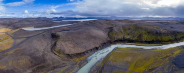 İzlanda 'nın engebeli volkanik arazisinin hava perspektifi, dolambaçlı bir nehir, yeşil yosun yamaları, uzak su ve bulutlu bir gökyüzünün altında yuvarlanan tepeler içerir..