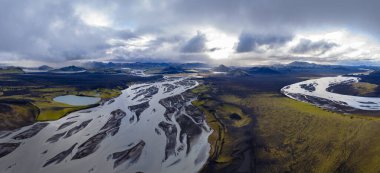 İzlanda 'nın örülmüş nehirlerinin karanlık volkanik kumlar üzerindeki hava perspektifi, yosun kaplı arazi, küçük bir göl ve bulutlu bir gökyüzünün altındaki uzak dağlar..