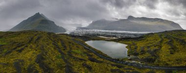 Bir buzul, bulutlu bir gökyüzünün altında volkanik dağlarla kaplı yosunlardan uzanıyor. Küçük bir yansıtıcı göl ve dolambaçlı yolları olan karanlık bir arazi görünür..