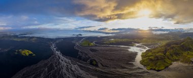 Karanlık volkanik ovaları aşan örülmüş nehirlerin hava perspektifi, yeşil tepeler, uzakta bir buzullar ve İzlanda 'da güneşli bir gökyüzü..