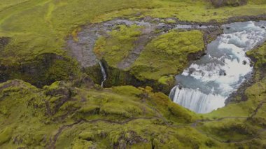 Yeşil İzlanda manzarası. Mavi bir nehre akan bir şelale. Etrafı yosun kaplı kayalar ve dolambaçlı patikalarla çevrili..