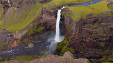Yüksek bir şelalenin hareket halindeki hava manzarası, yükselen sisli bir kayalık havza, etrafını saran uçurumlar, yosunlar, volkanik kayalar ve dönen bir nehir..