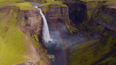 Uzun bir şelale yeşil kayalıklar ve volkanik kayalarla çevrili derin bir kanyona akar. Bir nehir engebeli arazide süzülürken sis yükselir..