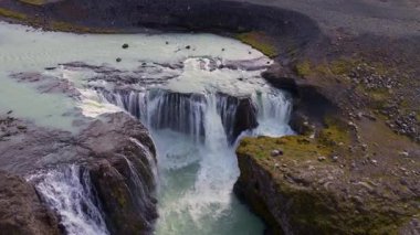 İzlanda 'da volkanik kayaların üzerinden akan turkuaz su yosun kaplı arazi ve dinamik hareketle çevrili bir şelale manzarası..